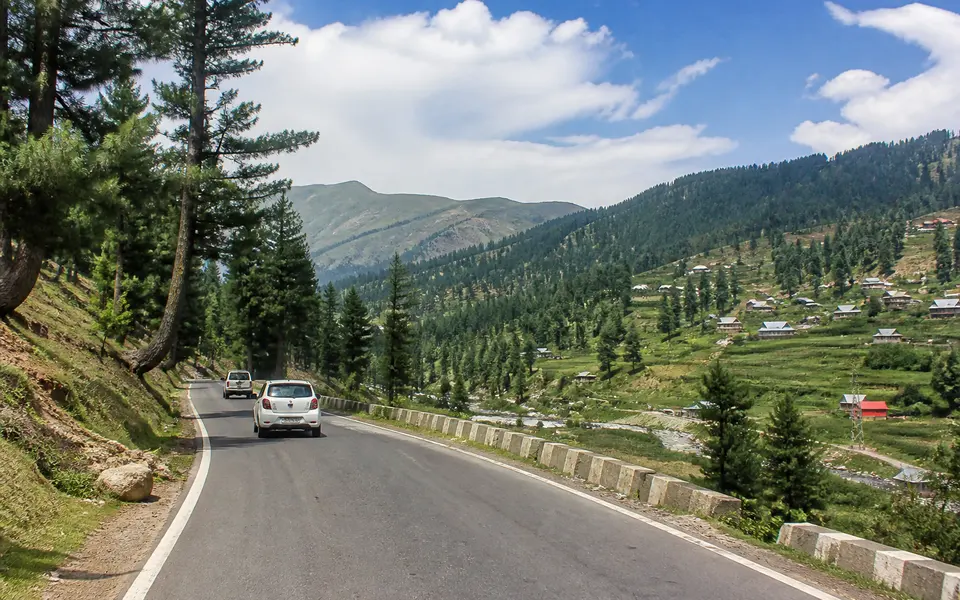 Paved hill road in Kashmir with cars driving through pine forest towards distant meadows.