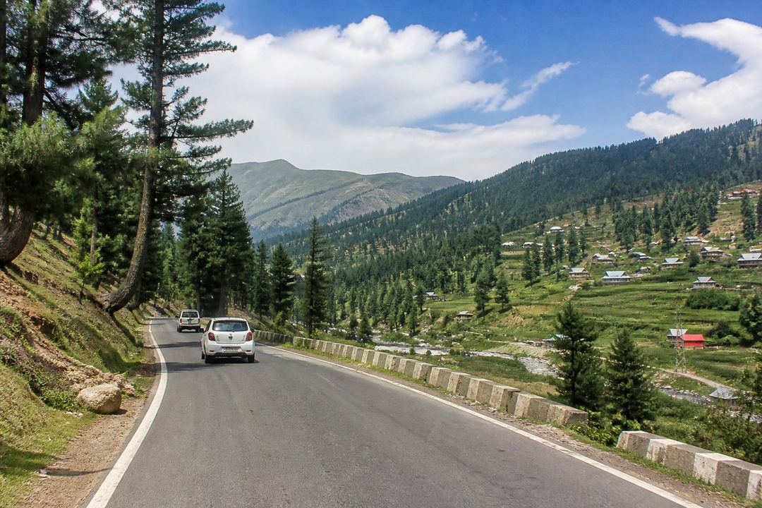 Paved hill road in Kashmir with cars driving through pine forest towards distant meadows.
