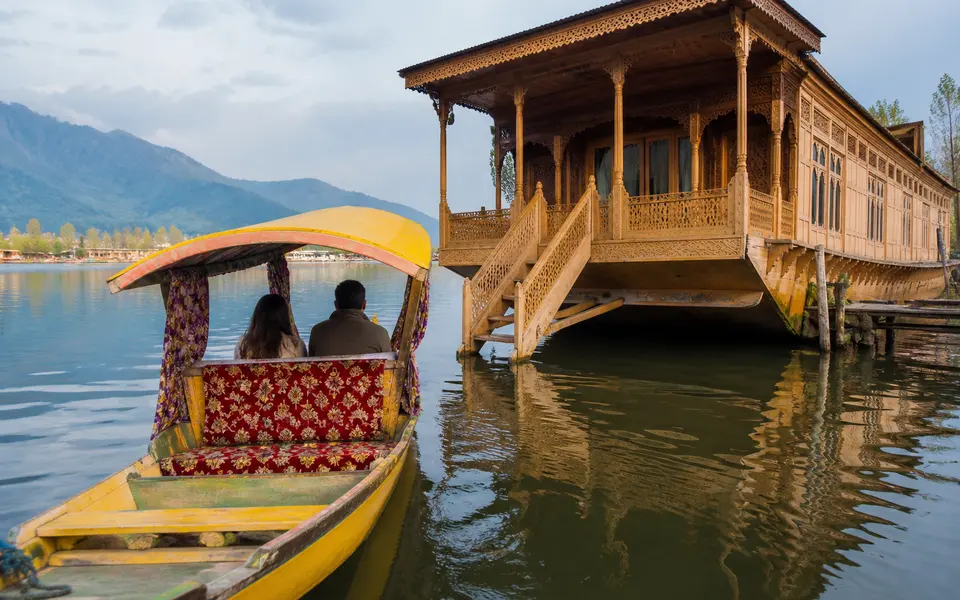 A shikara boat approaching a traditional wooden houseboat on Dal Lake, with guests seated under a canopy.