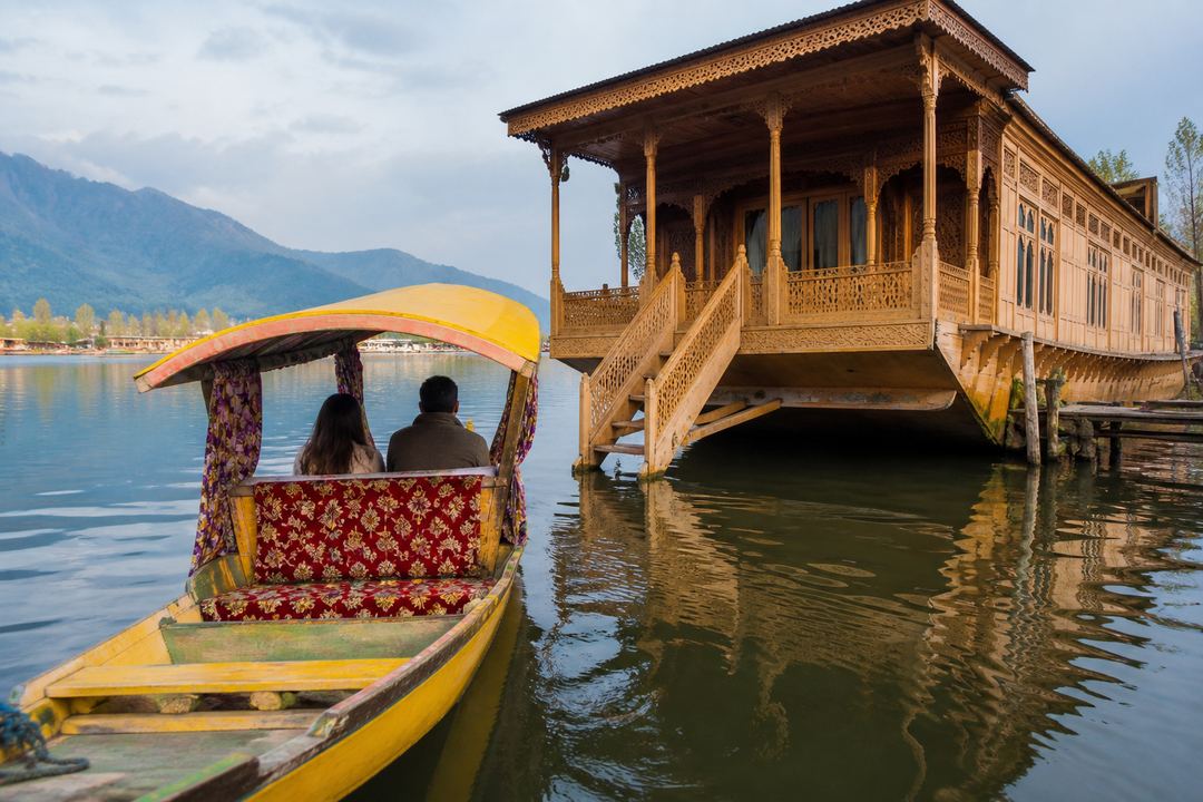 A shikara boat approaching a traditional wooden houseboat on Dal Lake, with guests seated under a canopy.