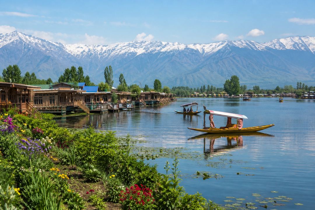 Dal Lake in Srinagar with houseboats and spring greenery in the foreground and snow still visible on the higher mountains in the background.
