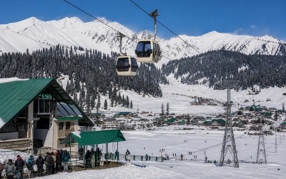 Cable cars of the Gulmarg Gondola rising above snow-covered Gulmarg town towards forested slopes and distant Himalayan peaks.