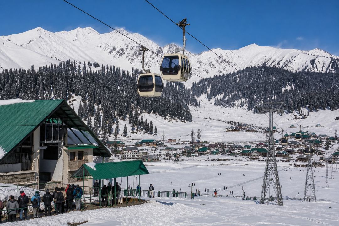 Cable cars of the Gulmarg Gondola rising above snow-covered Gulmarg town towards forested slopes and distant Himalayan peaks.