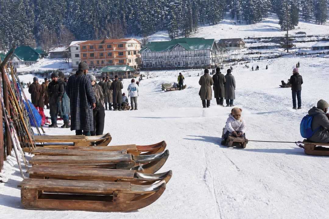 Families playing in the snow and using sledges on a gentle slope near Gulmarg’s base, with pine trees and low buildings in the background.