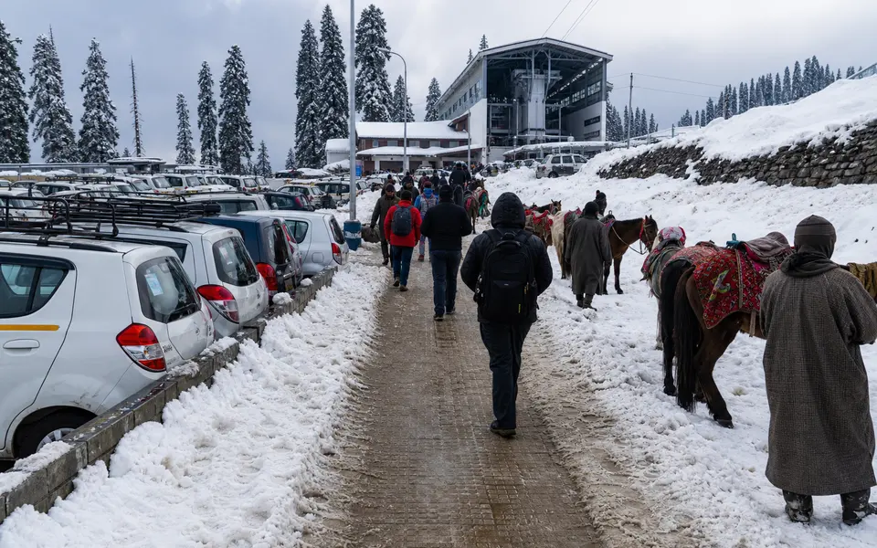 Approach road and pedestrian path to the Gulmarg Gondola base station with snow, parked vehicles, ponies and visitors walking in winter clothing.