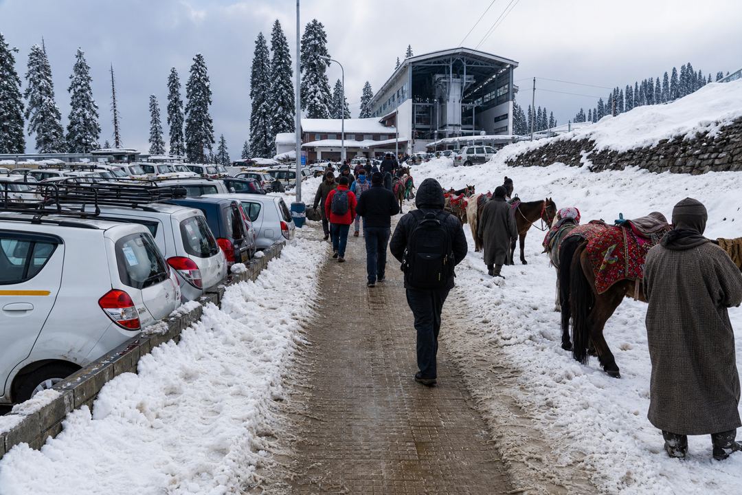 Approach road and pedestrian path to the Gulmarg Gondola base station with snow, parked vehicles, ponies and visitors walking in winter clothing.