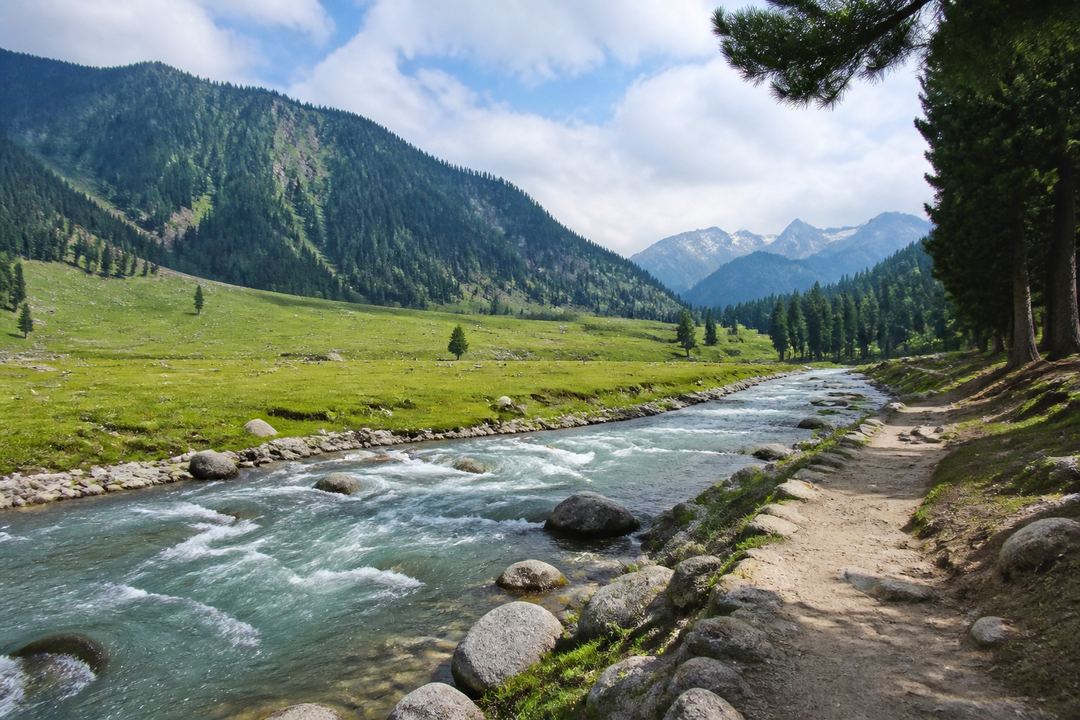 Calm stretch of the Sindh River near Sonamarg with a riverside path, flat meadow and distant mountains.