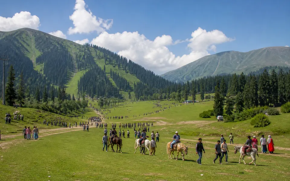 Summer view of Gulmarg meadow with green slopes, pine trees and visitors walking and riding horses under a bright sky.