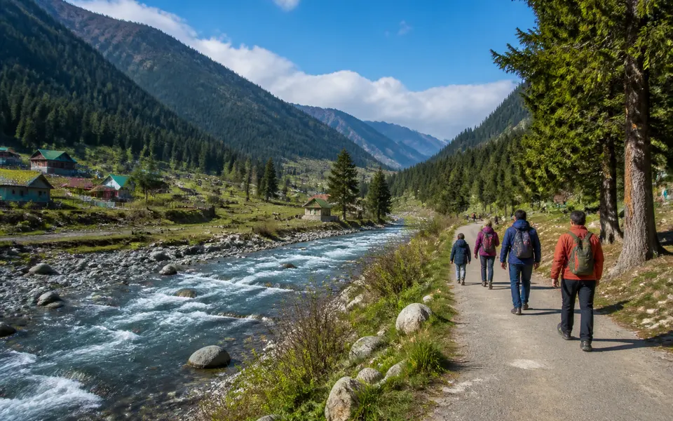 Wide view of Pahalgam valley with a river, pine forests and open meadows, with a few travellers walking along the riverside.