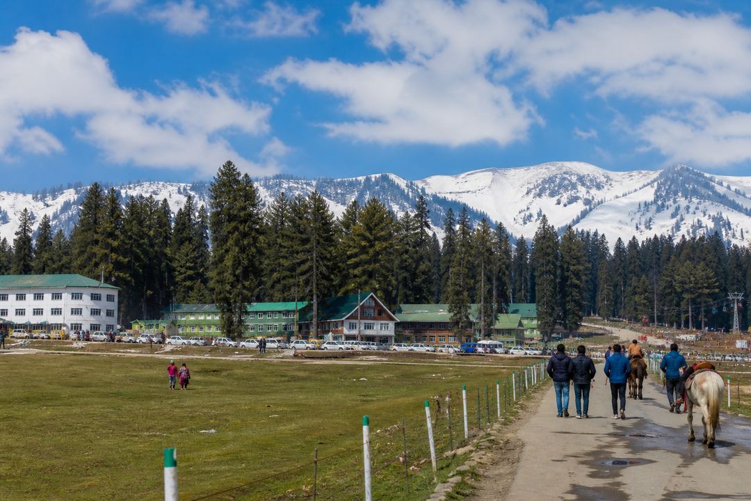 Ground-level view of Gulmarg’s central meadow with pine trees, hotels and visitors walking along the snow or grass.