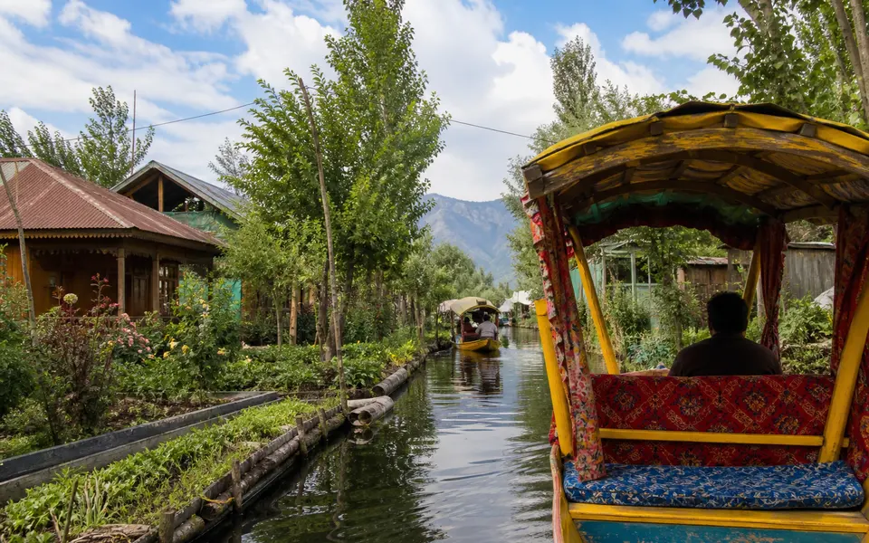 A shikara moving through a narrow canal of Dal Lake lined with floating gardens and small houses.