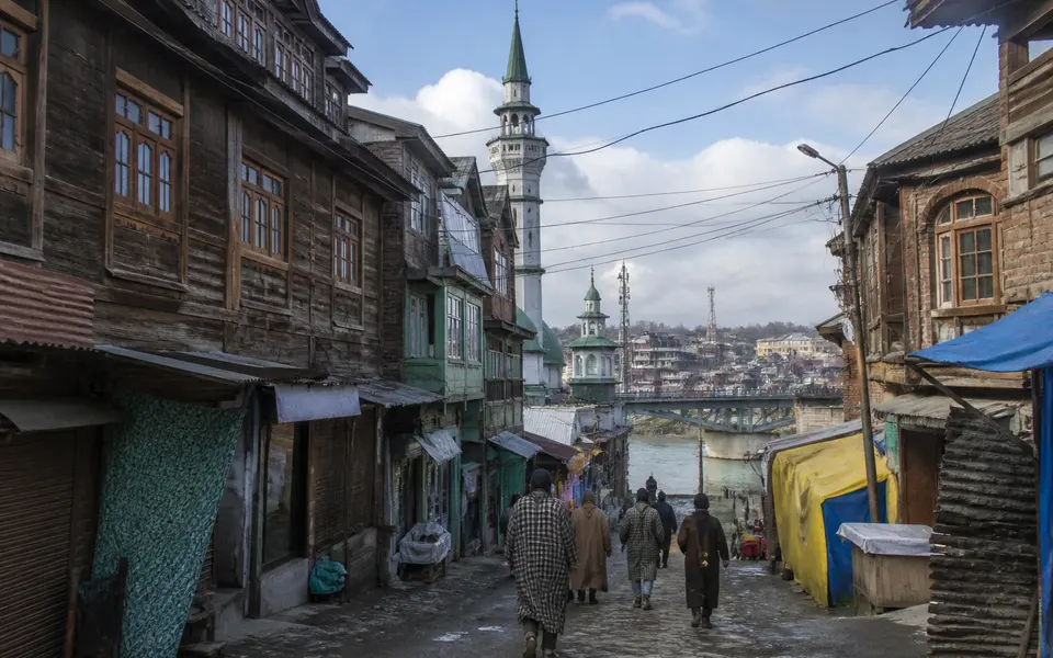 Narrow street in Srinagar’s Old City with traditional timber-fronted buildings, a mosque minaret and people walking towards the Jhelum river.