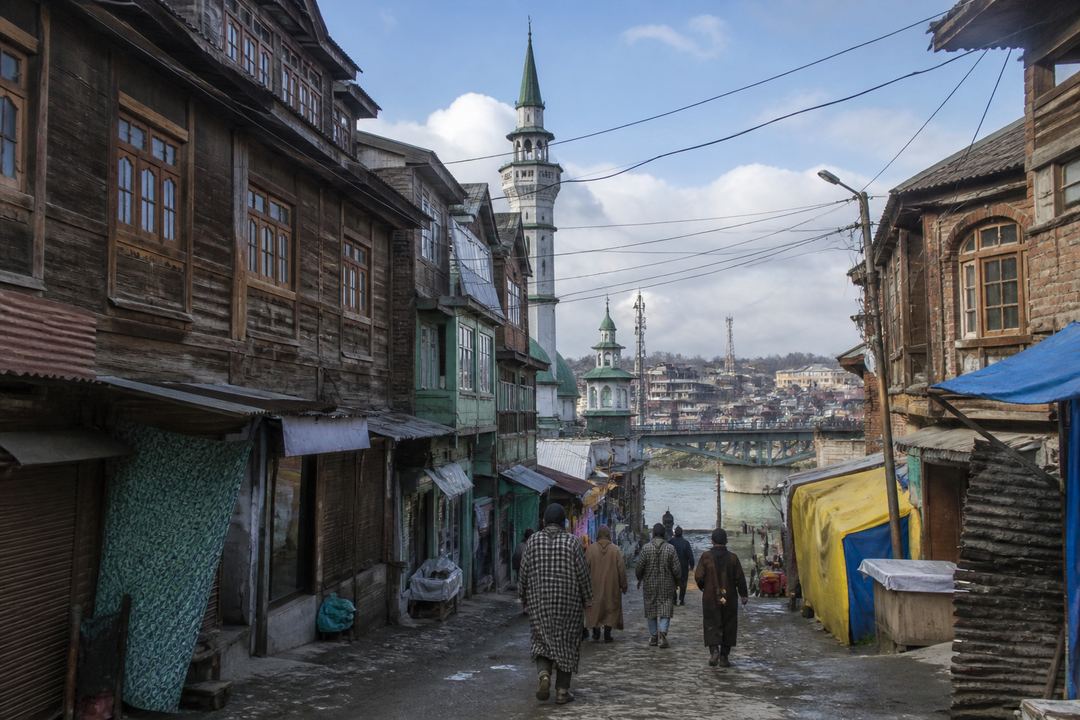 Narrow street in Srinagar’s Old City with traditional timber-fronted buildings, a mosque minaret and people walking towards the Jhelum river.