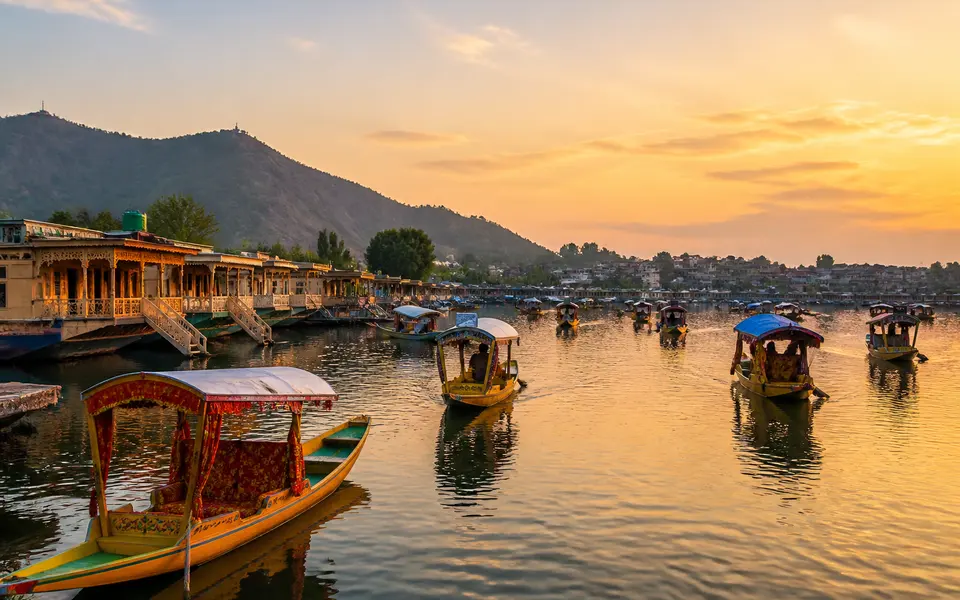Shikaras and houseboats on Dal Lake at sunset with the Srinagar city shoreline and hills in the background.