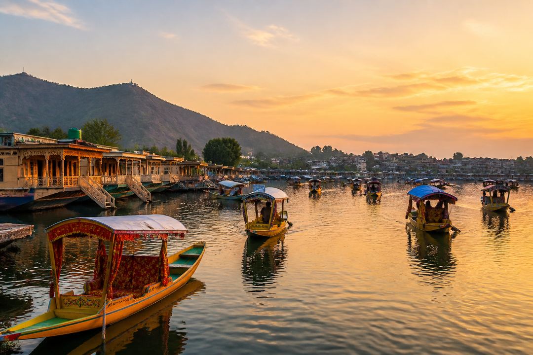 Shikaras and houseboats on Dal Lake at sunset with the Srinagar city shoreline and hills in the background.