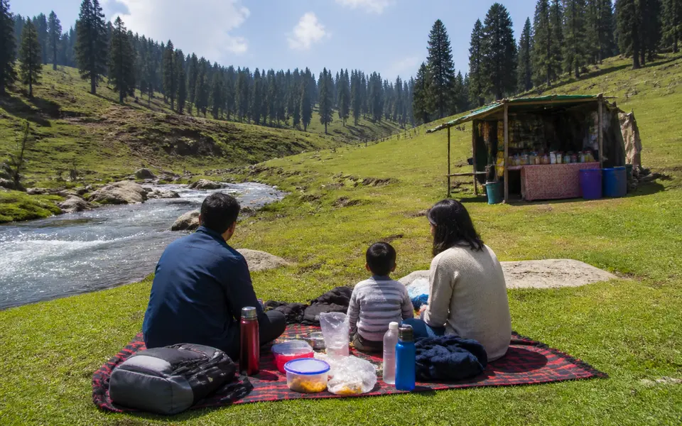 Family sitting on a picnic mat near a stream in a high meadow, with a small tea stall in the background.