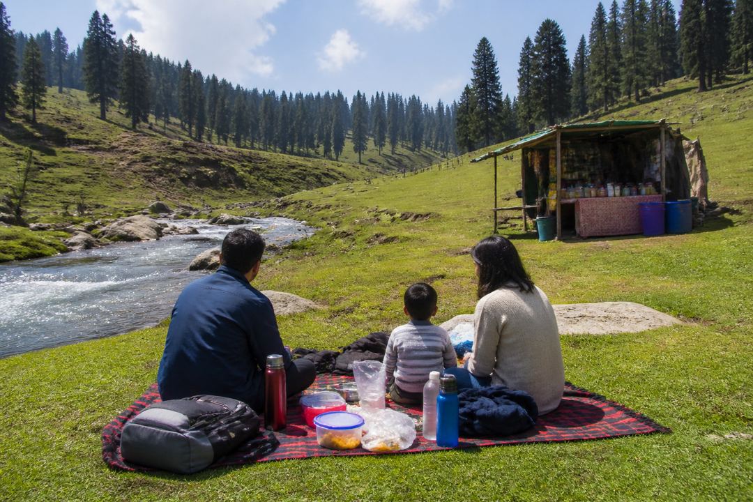 Family sitting on a picnic mat near a stream in a high meadow, with a small tea stall in the background.