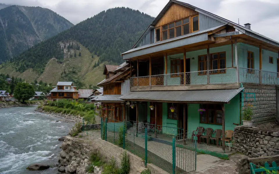 Small guesthouse or homestay in Dawar village overlooking the Kishanganga River, with wooden houses and mountains nearby.