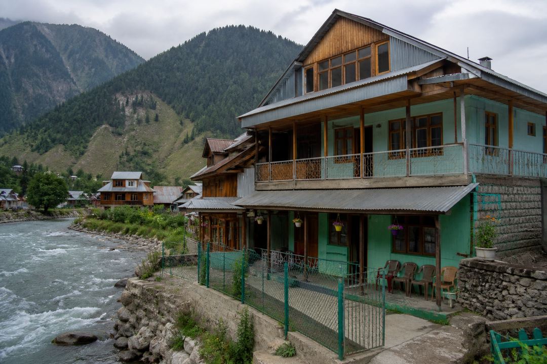 Small guesthouse or homestay in Dawar village overlooking the Kishanganga River, with wooden houses and mountains nearby.