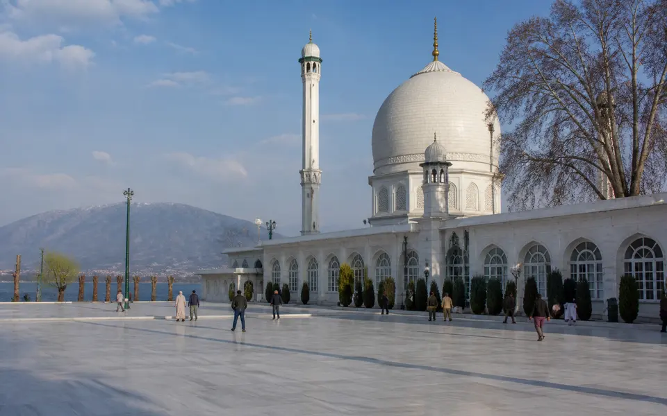 Courtyard of Hazratbal Shrine with its white domes beside the lake, visitors walking in the open space and hills in the background.