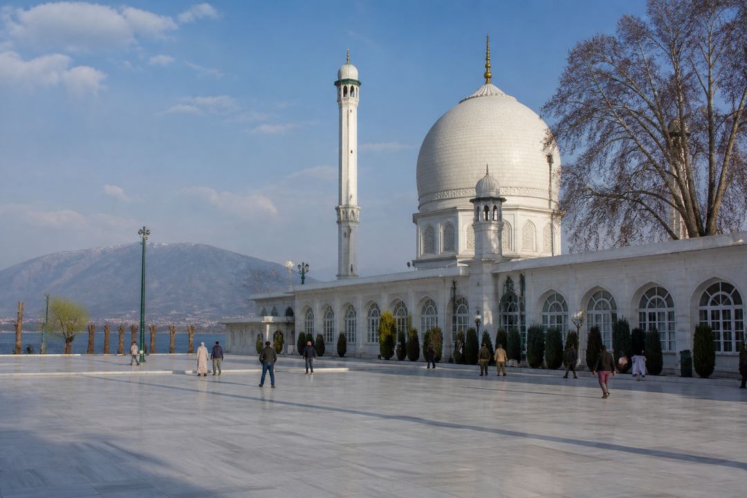 Courtyard of Hazratbal Shrine with its white domes beside the lake, visitors walking in the open space and hills in the background.