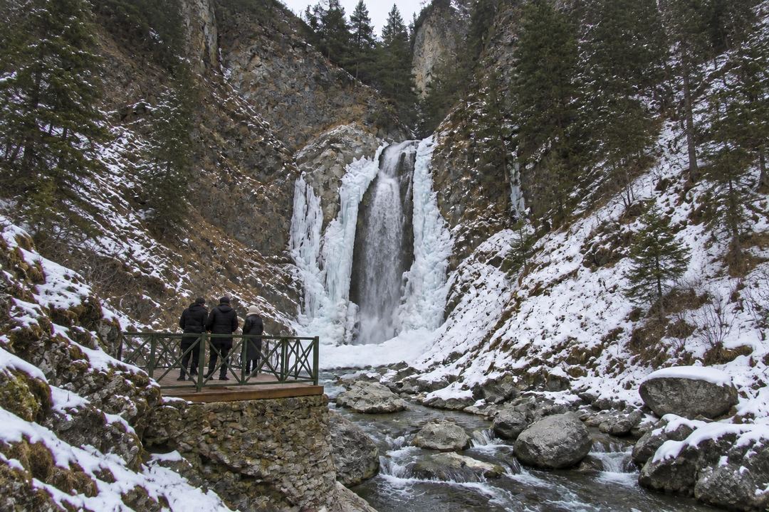 Waterfall in a rocky Himalayan valley near Gulmarg, partly frozen in winter with snow patches and pine trees on the slopes.